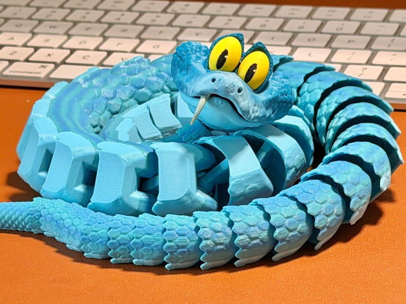 May include: A coiled, articulated blue snake toy with a detailed, textured design. The snake features large yellow eyes with black pupils and a forked tongue. The toy is made of plastic and sits on an orange surface.
