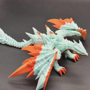 Op de afbeelding: Gedetailleerd beeldje van een draak, met een lichtblauw lichaam en oranje accenten op de vleugels, kop en klauwen. De draak heeft een lange staart en staat op een donkere ondergrond. Het beeldje is een verzamelobject met fantasiethema.