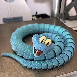 May include: A coiled, articulated snake toy in shades of blue. The snake has a detailed, textured body and a smiling head with large, yellow eyes and a forked tongue. The toy is likely 3D-printed and sits on a light brown surface.