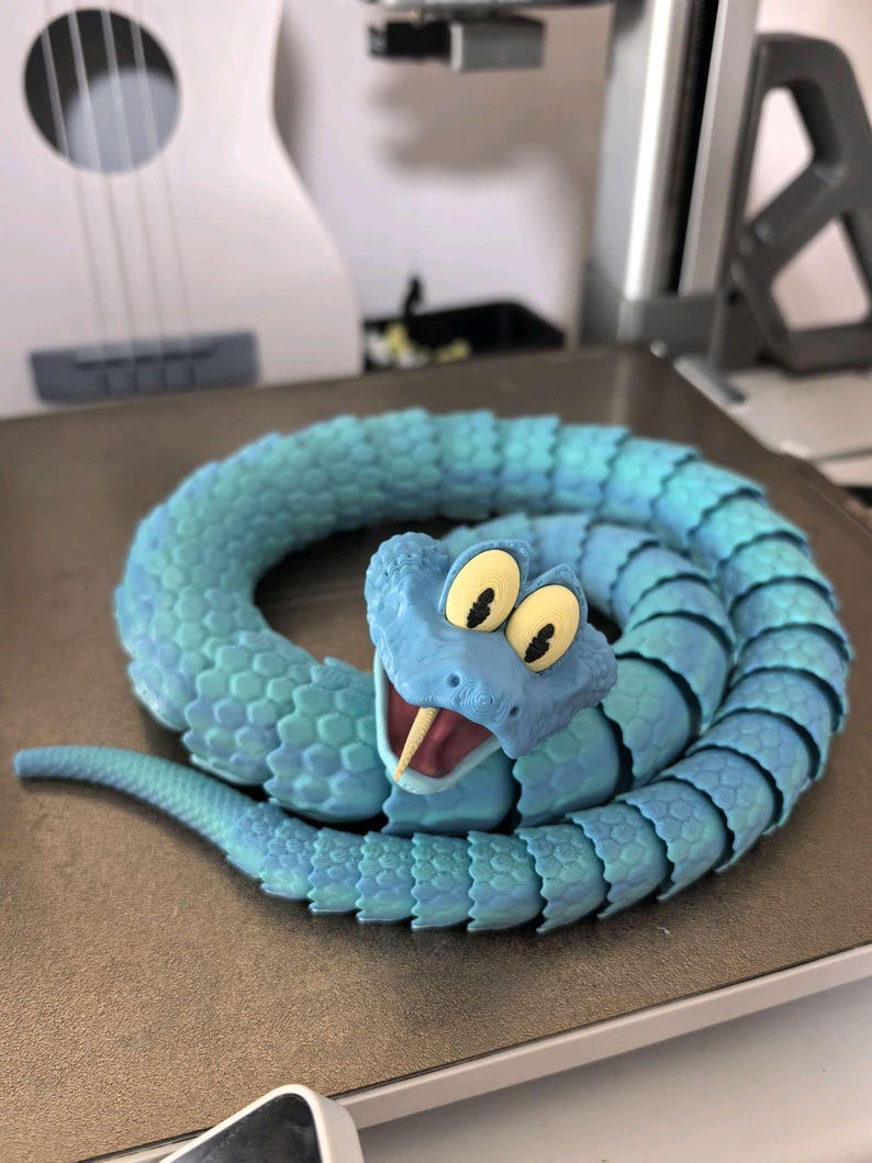 May include: A coiled, articulated snake toy in shades of blue. The snake has a detailed, textured body and a smiling head with large, yellow eyes and a forked tongue. The toy is likely 3D-printed and sits on a light brown surface.