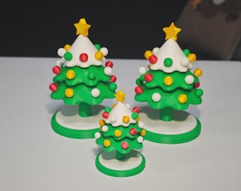 Puede incluir: Tres adornos navideños en miniatura con forma de árbol de Navidad. Cada árbol tiene una base y un cuerpo verdes, una parte superior blanca y está adornado con adornos coloridos y una estrella amarilla. Los árboles varían en tamaño, el más grande mide unos 10 cm de alto.
