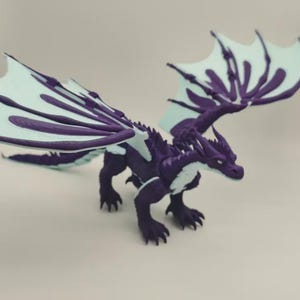 Op de afbeelding: Een paars en wit drakenbeeldje. De draak heeft een donkerpaars lichaam met witte accenten en lichtpaarse vleugels. De vleugels zijn gespreid en de draak staat in een looppositie. Fantasiewezen.