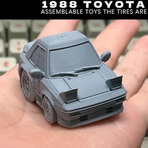 Op de afbeelding: Een grijze 1988 Toyota MR2 speelgoedauto met beweegbare banden, vastgehouden in een hand. De auto heeft pop-up koplampen en zijspiegels. Tekst bovenaan luidt "1988 TOYOTA MR2 ASSEMBLABLE TOYS THE TIRES ARE MOVABLE!"