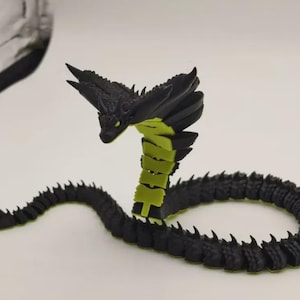 Può includere: Una statuetta di drago articolata nera e verde. Il drago ha una coda spinosa dettagliata e una testa con una struttura a forma di ala svasata. Il corpo segmentato è nero con accenti verdi e gli occhi sono gialli.