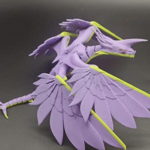 Könnte beinhalten: Detaillierte, lila Drachenfigur mit gelben Akzenten auf Flügeln und Körper. Der Drache hat einen langen Hals, einen detaillierten Kopf und ausgebreitete Flügel. Die Figur steht vor einem grauen Hintergrund.