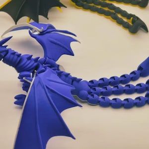 Op de afbeelding: Een blauw gearticuleerd drakenbeeldje met grote vleugels en een gesegmenteerde staart. De draak heeft een gedetailleerd ontwerp met stekels langs de rug en een gebogen hoorn. Een andere draak met gele en zwarte vleugels is zichtbaar.