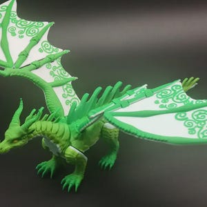 Op de afbeelding: Een groene drakenfiguur met wit en groen patroonvleugels. De draak heeft een gedetailleerd ontwerp, met een lange nek, hoorns en scherpe klauwen. De vleugels zijn uitgespreid en tonen de ingewikkelde patronen.