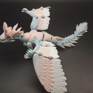 Op de afbeelding: Een gedetailleerd, gearticuleerd drakenbeeldje in pastelkleuren. De draak heeft een lichtblauw lichaam, roze accenten en grote gevederde vleugels. De staart en kop hebben ingewikkelde details en rode ogen.
