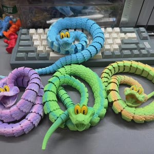 May include: Four colorful, articulated snake toys in shades of blue, green, yellow, and purple. Each snake has a smiling face with large yellow eyes. The toys are coiled on a gray surface.