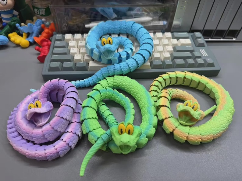 May include: Four colorful, articulated snake toys in shades of blue, green, yellow, and purple. Each snake has a smiling face with large yellow eyes. The toys are coiled on a gray surface.