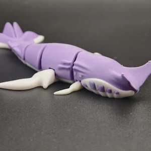 Könnte beinhalten: Ein lila-weißes, bewegliches Walspielzeug. Das Spielzeug hat eine hellviolette Farbe mit weißen Akzenten an den Flossen und am Bauch. Der Wal liegt auf einer dunklen Oberfläche.