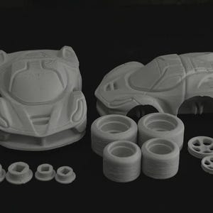 Op de afbeelding: Een verzameling onbeschilderde, lichtgrijze 3D-geprinte auto-onderdelen, waaronder twee carrosserieën, vier banden, vier wielen en diverse kleinere componenten. De onderdelen zijn op een donkere ondergrond geplaatst.