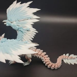 Peut inclure: Figurine détaillée d'un dragon avec de grandes ailes en plumes. Le dragon est principalement blanc et bleu clair, avec une queue segmentée dans des tons de rose et de blanc. La figurine est posée sur une surface sombre.