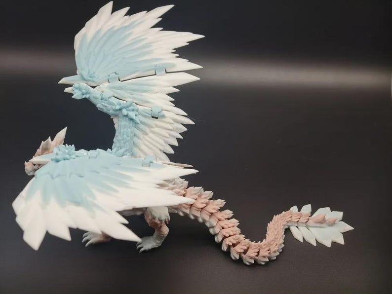 May include: A detailed figurine of a dragon with large, feathered wings. The dragon is primarily white and light blue, with a segmented tail in shades of pink and white. The figure is posed on a dark surface.