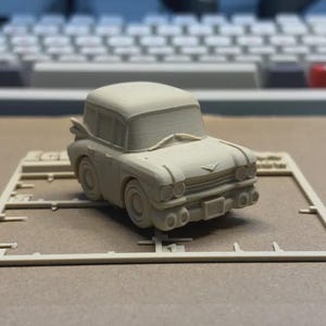 May include: A beige model car with a classic design, featuring rounded headlights and a prominent front grille. The car is on a beige plastic frame with measurement markings, suggesting it's a model kit. The background includes a keyboard.