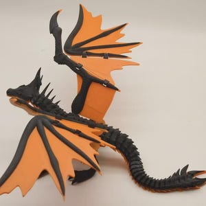 Op de afbeelding: Een 3D-geprinte drakenfiguur met oranje vleugels en zwarte accenten. De draak heeft een gedetailleerde, stekelige rug en staart, met een zwarte kop en hoorns. De vleugels zijn uitgespreid.
