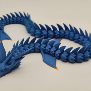 May include: A blue articulated dragon toy with a detailed design. The dragon has a head, body, and tail, all in a vibrant blue color. The dragon's head has orange accents, and the tail has a fin-like shape.