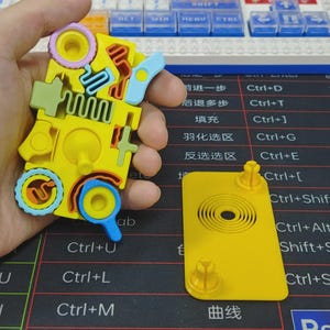 May include: A yellow puzzle toy with various colorful shapes and cutouts, held in a hand. The puzzle has blue, green, pink, and orange elements. A separate yellow piece with a circular design and pegs is also visible. The background includes a keyboard.