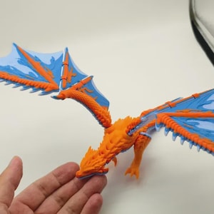 May include: An orange and blue dragon figurine with detailed features and outstretched wings. The wings are blue with orange detailing, and the body is orange. The dragon is posed as if in flight, with its wings spread wide.