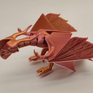 Könnte beinhalten: Eine 3D-gedruckte Drachenfigur in Rosa- und Orangetönen. Der Drache hat detaillierte Flügel, Hörner und Krallen. Die Figur ist mit ausgebreiteten Flügeln posiert und zeigt das komplizierte Design und die Textur.