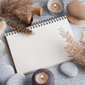 May include: A spiral-bound notebook with blank white pages, surrounded by dried beige grasses, smooth gray and tan stones, and two lit candles in small containers. The arrangement is set against a weathered white wood-grain surface.