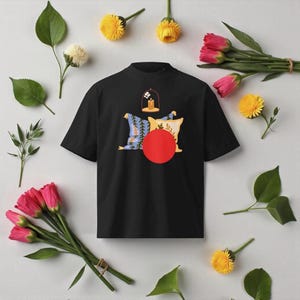 Może przedstawiać: Czarny t-shirt z krótkim rękawem i wysokim dekoltem. Koszulka posiada grafikę świecy w szklanej kopule, poduszek i czerwonego koła. Obraz otoczony jest kwiatami i liśćmi.