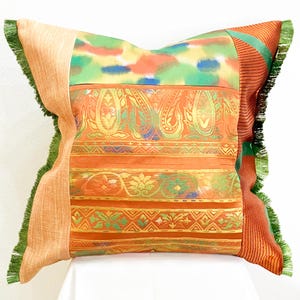 May include: A square throw pillow featuring a patchwork design of orange, green, and multicoloured fabric panels. The pillow includes sections with paisley and floral patterns, accented by a solid light orange side panel and green fringe trim.
