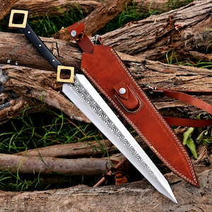 May include: A double-edged sword with a black handle, gold-toned square pommel and guard, and a silver blade featuring a repeating fan-shaped pattern. The sword is accompanied by a brown leather sheath with a snap closure and adjustable strap.