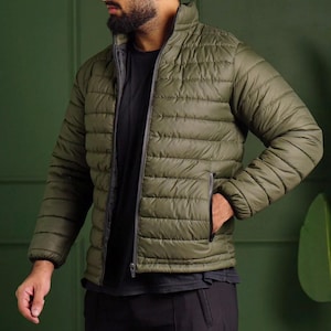 Könnte beinhalten: Olivgrüne Steppjacke mit schwarzem Reißverschluss und zwei Seitentaschen. Die Jacke hat einen Stehkragen und ein dunkelgraues Futter. Ein leichtes, lässiges Oberbekleidungsstück.