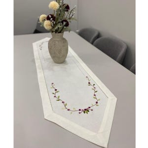 Puede incluir: Camino de mesa blanco bordado con un diseño floral. El camino de mesa presenta un delicado patrón de hojas verdes y bayas moradas. Un jarrón beige con flores se encuentra sobre el camino de mesa, agregando elegancia.
