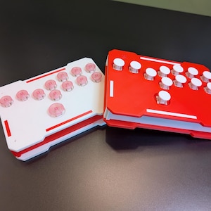 May include: Two arcade fight sticks, one white and one red, are displayed. Each controller has a rectangular design with multiple buttons. The white controller has pink buttons, while the red controller has white buttons. Both controllers have a sleek, modern aesthetic.