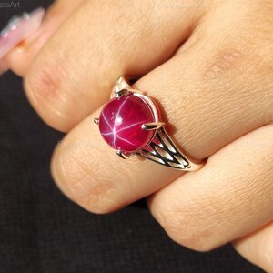 May include: A silver ring featuring a large, round, star ruby gemstone. The ruby is a deep red color with a visible star pattern. The ring has a delicate, openwork design on the band.