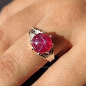 Könnte beinhalten: Ein silberner Ring mit einem großen, runden Sternrubin-Edelstein. Der Rubin zeigt einen sechsstrahligen Sterneffekt. Der Ring wird an einer Hand getragen, um das Design und die Farbe des Schmucks hervorzuheben.