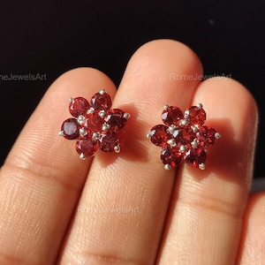 May include: A pair of flower-shaped stud earrings. Each earring features a cluster of small, round, deep red gemstones set in a silver-colored metal. The earrings are displayed against a dark background.