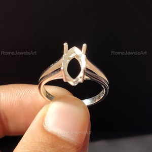 May include: A silver ring setting with an oval-shaped center and two prongs. The ring is held by a hand against a dark background. The ring is designed to hold a gemstone.