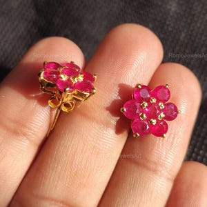 Op de afbeelding: Een paar bloemvormige oorstekers. Elke oorbel heeft een cluster van levendige rode edelstenen gezet in goudkleurig metaal. De oorbellen zijn ontworpen om op een bloem met meerdere bloemblaadjes te lijken.