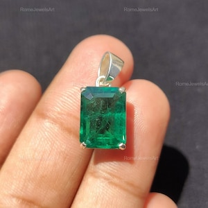 Könnte beinhalten: Ein rechteckiger, smaragdgeschliffener grüner Edelsteinanhänger in einer silberfarbenen Metallfassung mit einer dreieckigen Öse an der Oberseite. Der Edelstein zeigt einen tiefen, lebendigen Grünton mit sichtbaren Einschlüssen.