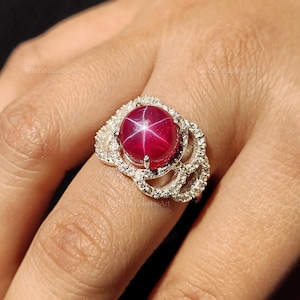 May include: A silver ring with a large, round, star ruby gemstone. The ruby is a deep red colour with a six-ray star effect. The ring has a detailed, openwork design with small, clear gemstones.