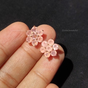 Puede incluir: Pendientes de botón con forma de flor en oro rosa. Cada pendiente presenta un grupo de piedras preciosas de color rosa claro dispuestas en un patrón floral. Fondo oscuro.