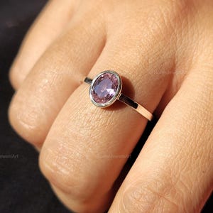 Könnte beinhalten: Ein silberner Ring mit einem ovalen, hellvioletten Edelstein in einer Zargenfassung. Der Ring befindet sich an einem Finger, mit einem dünnen, polierten Band. Der Edelstein ist der Fokus und reflektiert das Licht.