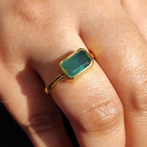 Peut inclure: Une bague en or ornée d'une pierre verte rectangulaire taille émeraude. La bague est sertie d'un simple anneau en or, mettant en valeur la couleur verte vibrante de la pierre. La bague est portée sur une main.
