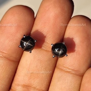 May include: A pair of black star sapphire stud earrings. Each earring has a round, polished black gemstone with a star pattern. The gemstones are set in a silver-coloured metal setting with four prongs.