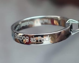 May include: A polished silver-coloured metal ring featuring the engraved text "Strong as Fuck" on the outer band. The ring is held by a small metal tool, and additional faint, partially visible text is engraved on the inner surface of the band.