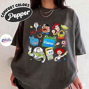 May include: Dark grey t-shirt featuring colorful cartoon characters from the Toy Story movie. The shirt includes images of Woody, Buzz Lightyear, and other characters. The text "TOY STORY" is also visible. The shirt has the text "COMFORT COLORS Pepper" on the top left.