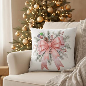 Puede incluir: Un cojín cuadrado blanco con un diseño de acuarela de un lazo rosa, agujas de pino, bayas y adornos. El cojín está sobre un sillón beige con un árbol de Navidad al fondo.