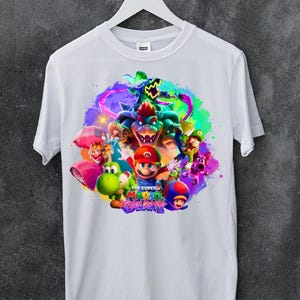 Puede incluir: Una camiseta blanca de manga corta con un estampado gráfico vibrante y colorido de Mario, Luigi, la Princesa Peach, Yoshi, Bowser y otros personajes de la serie de videojuegos The Super Mario Galaxy, centrado en el pecho con un fondo salpicado.
