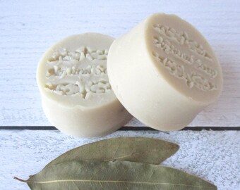 May include: Two round, light beige bars of soap with a stamped decorative pattern and text on the top surface. The soap bars are positioned behind two dried, elongated green bay leaves. The texture of the soap appears smooth and solid.