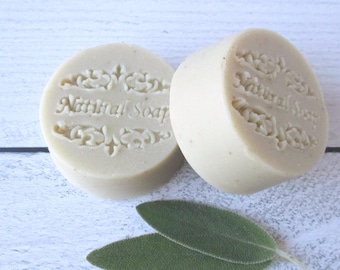 May include: Two round, beige-colored bars of soap with a smooth texture. Each bar features an embossed decorative border and the text "Natural Soap" centered on the top surface. Two green sage leaves are positioned below the soap bars.