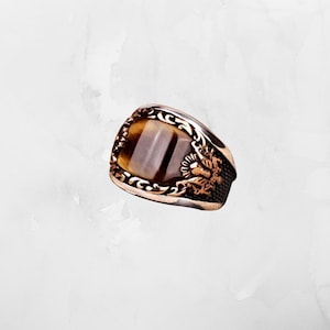 May include: A bronze ring with a rectangular tiger's eye stone. The ring has ornate detailing around the stone and a dragon motif on the side. The stone is a mix of brown and tan colours.