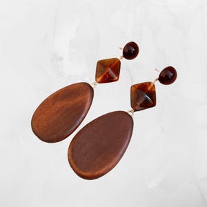 Puede incluir: Un par de pendientes colgantes con elementos de madera marrón oscuro, cuadrados y redondos de color ámbar y alambres de conexión dorados. Los pendientes tienen un cierre de tipo poste.